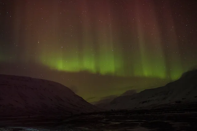 Curtains of green and red northern lights rising above dark, snow-covered mountains under a starry sky
