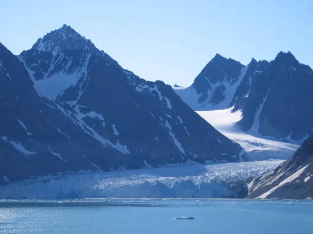 A glacier flowing between steep, snow-covered peaks into the blue water of Magdalenefjorden under a clear sky
