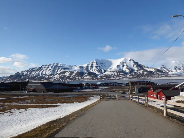 A paved road with a wooden fence leading down towards the houses of Longyearbyen, with snowy mountains across the fjord under a blue sky